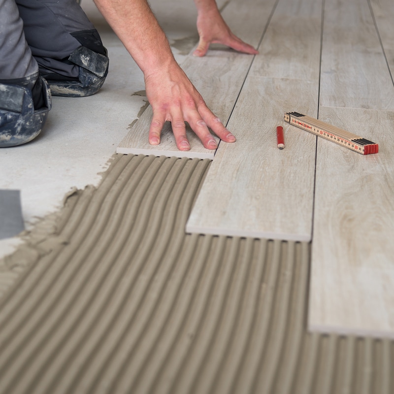 Mains posant des carreaux imitation bois sur adhésif crénelé. Sur le sol, une règle pliante et un crayon rouge sont visibles.