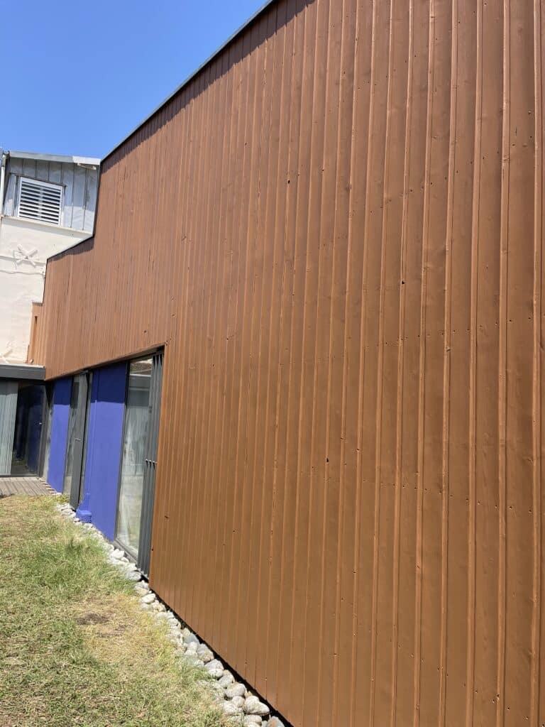 Bardage bois moderne brun: façade contemporaine, accents bleus Façade de bâtiment moderne avec bardage bois brun vertical, sections bleues vives, grandes fenêtres et base de galets sous ciel azur.