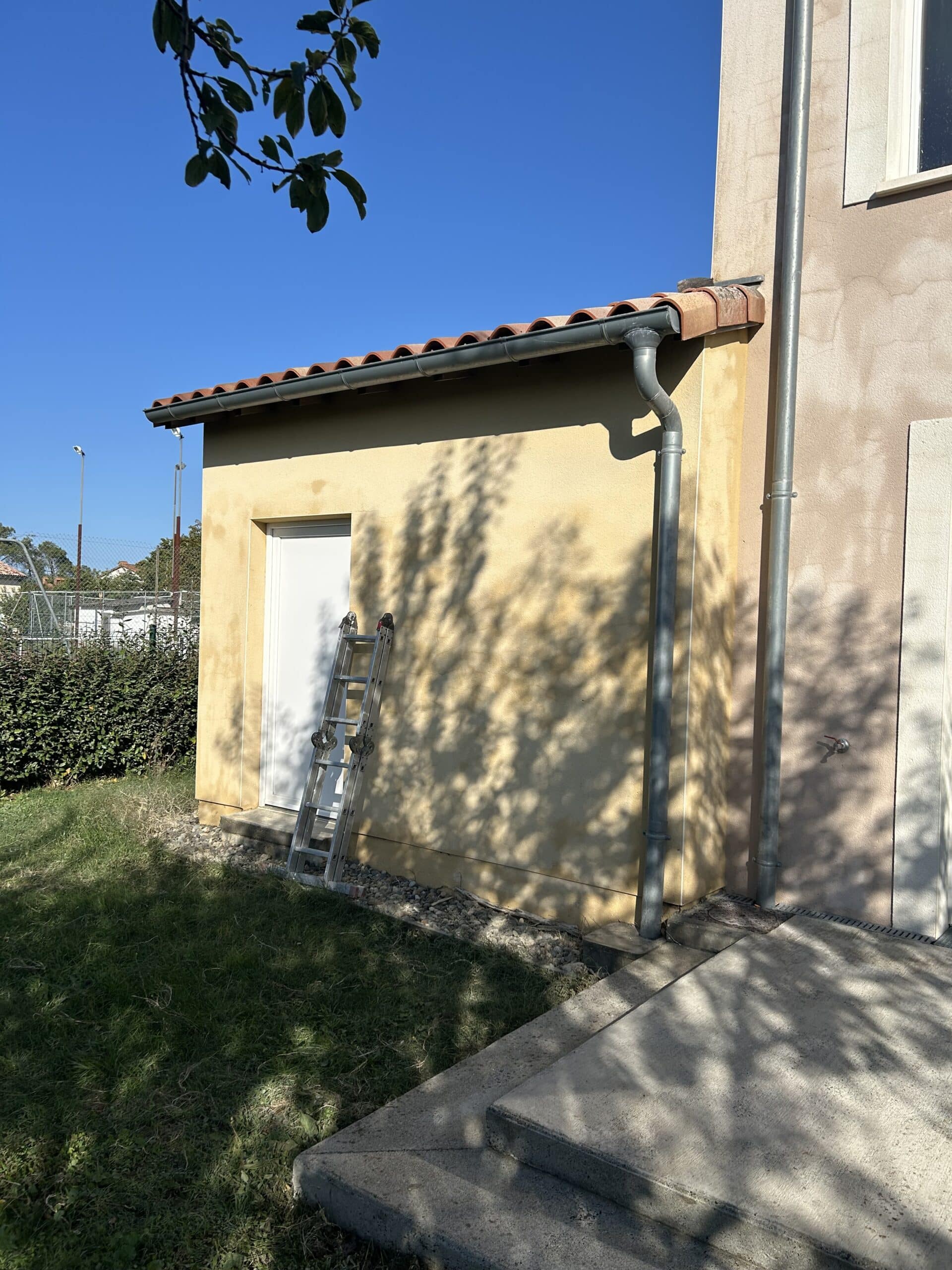 Annexe jaune ensoleillée avec échelle, toiture tuilée, gouttière, et un robinet extérieur. Ombre d'arbre sur le mur et la terrasse en béton.