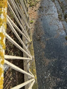 Balustrade métallique moussue en surplomb d'un sol herbeux et d'une surface asphaltée mouillée, parsemée de débris et de taches blanches.