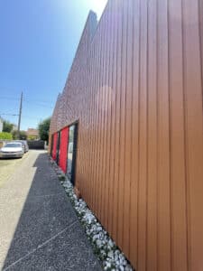 Façade moderne en lattes de bois marron-rouge d'un bâtiment angulaire, avec fenêtres rouges vives, sous un ciel bleu clair. Voiture VW FJ-311-XP garée à gauche.