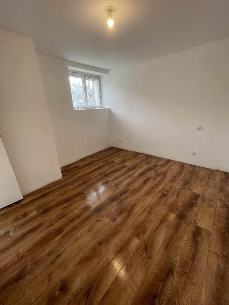Chambre rénovée : murs blancs, parquet stratifié moderne Chambre rénovée vide aux murs blancs, parquet stratifié marron-orangé brillant, petite fenêtre et prises électriques murales.