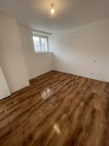 Chambre vide rénovée avec murs blancs et parquet stratifié marron brillant, une fenêtre lumineuse et un plafonnier simple.