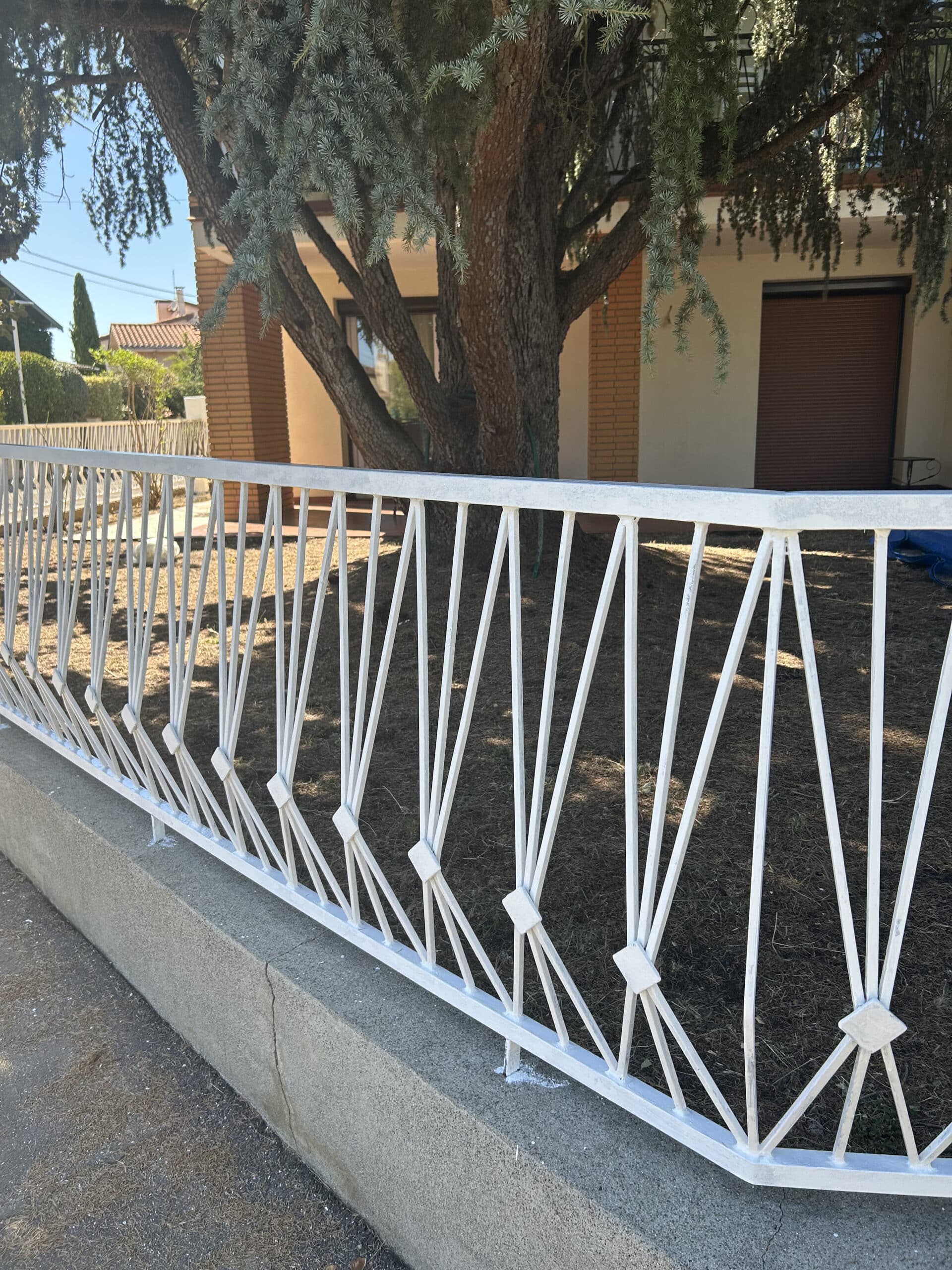 Clôture métallique blanche à motifs géométriques devant un grand cèdre, un jardin sec et une maison avec volet marron.