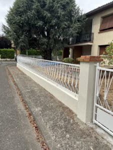 Clôture métallique blanche à motifs modernes devant un jardin avec grand cèdre, une maison beige et un trottoir.