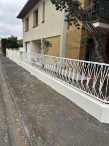 Maison beige et brique, avec clôture blanche fraîchement peinte à motif géométrique, le long d'un trottoir en béton. Arbre et haies.
