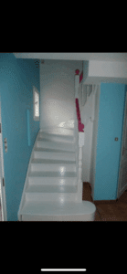 Escalier intérieur blanc avec main courante rose vif et murs turquoise lumineux. Une petite fenêtre et une porte blanche sont visibles.