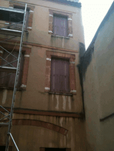 Façade beige d'un bâtiment ancien avec fenêtres aux volets violets fermés, encadrements de briques rouges. Traces d'humidité et échafaudage sur la gauche.
