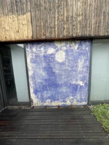 Façade architecturale avec bardage bois vertical clair et foncé, un grand mur central bleu vieilli et écaillé, flanqué de fenêtres sombres, et une terrasse en bois foncé au sol.