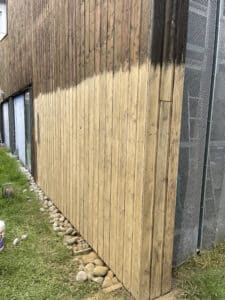 Façade de bâtiment en bois avec démarcation: haut foncé et vieilli, bas bois clair fraîchement nettoyé. Herbe, pierres, panneau perforé.