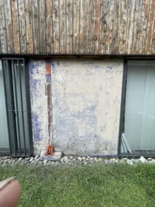 Bardage bois vieilli, mur bleu-blanc écaillé avec tuyau orange, fenêtres à barreaux, et sol en pierres/herbe.