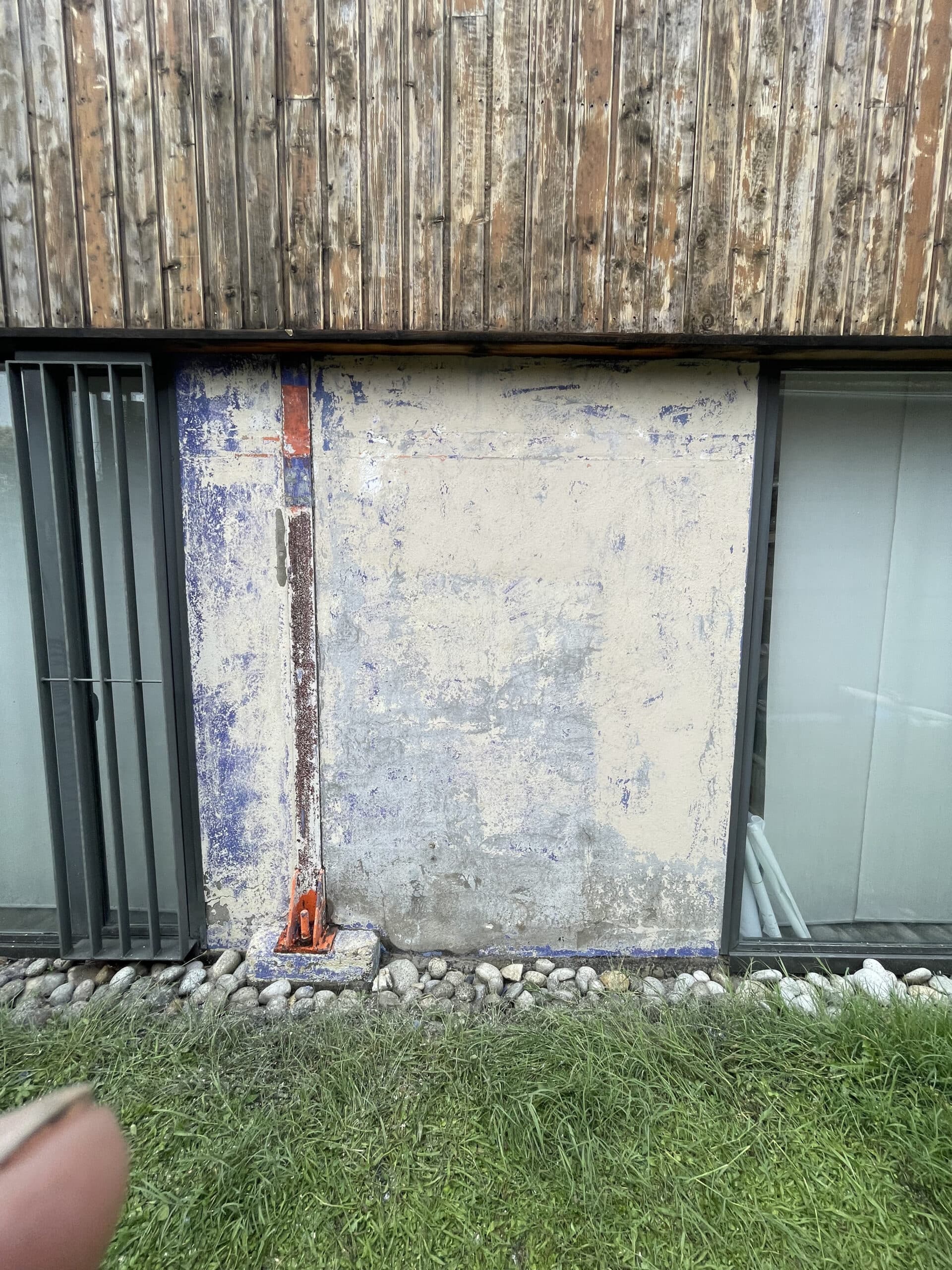 Bardage bois vieilli, mur bleu-blanc écaillé avec tuyau orange, fenêtres à barreaux, et sol en pierres/herbe.