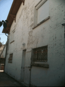 Façade de maison décrépie et fissurée. Toit en bois, volet roulant blanc, porte battante, et deux fenêtres à barreaux décoratifs.