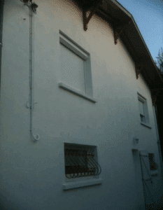 Façade blanche de maison. Fenêtres avec volet roulant et grilles en fer forgé. Gouttière et corniches en bois sombre.
