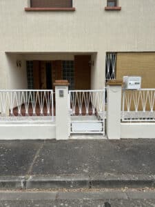 Façade maison beige, entrée avec porte bois et briques. Clôture et portillon blancs devant le numéro 25 et une boîte aux lettres.