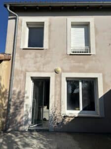 Façade maison beige deux étages, gouttière à gauche. Fenêtres (une avec store), porte-fenêtre ouverte, traces d'usure. Lumière murale, ombres d'arbres sur patio.