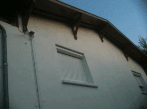 Façade de maison blanche avec volets roulants fermés, descente pluviale grise et avant-toit en bois.
