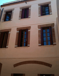 Façade d'un bâtiment couleur pêche avec six fenêtres à volets rouges bordeaux ouverts. Les cadres sont en briques et bois. Style méditerranéen.