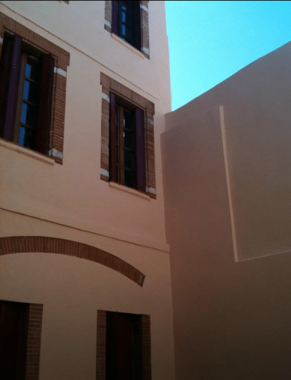 Façade rose pâle avec fenêtres à volets foncés, cadres en brique et grande arche. Mur minimaliste adjacent sous ciel bleu clair.