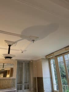 Intérieur Art Déco avec moulures de plafond blanches complexes, luminaire suspension stylisé, papier peint texturé et grandes fenêtres anciennes donnant sur un jardin verdoyant.
