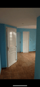 Intérieur d'une pièce lumineuse aux murs bleu turquoise et sol carrelé marron. Trois portes blanches, une ouverte à gauche.