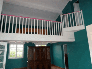 Intérieur coloré avec murs turquoise, mezzanine blanche et balustrade rose. Un grand buffet en bois est devant des fenêtres lumineuses.