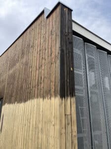 Façade en bois d'un bâtiment, partiellement nettoyée. Le bois clair ressort face au bois vieilli foncé, près de panneaux métalliques perforés.