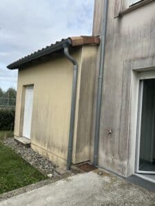 Façades beige et grise d'une maison avec des traces de moisissure et de saleté. Gouttières et descentes d'eau, porte blanche, robinet extérieur et dalles de drainage.
