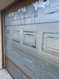 Porte de garage marron avec peinture grise fortement écaillée, révélant la rouille sur les panneaux et de nombreuses coulures d'eau.