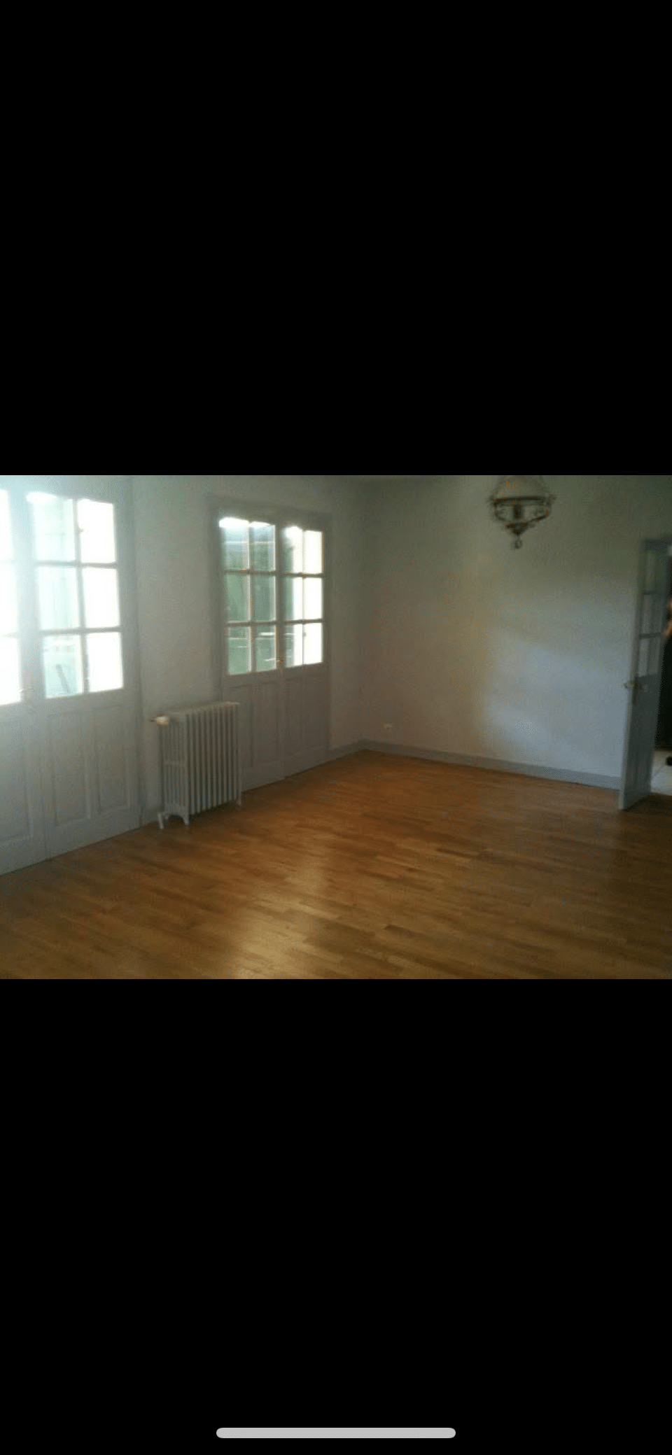Pièce vide lumineuse avec parquet en bois clair, deux grandes fenêtres, radiateur et un lustre au plafond.