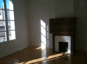 Pièce vide lumineuse avec parquet clair, murs blancs, grande fenêtre à carreaux. La lumière du soleil projette des ombres sur le sol et la cheminée.