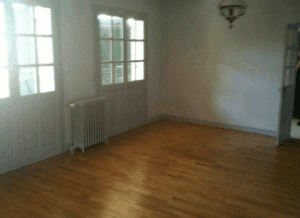 Pièce vide lumineuse. Parquet clair, deux grandes fenêtres-portes, radiateur blanc et porte ouverte. Lustre vintage au plafond.
