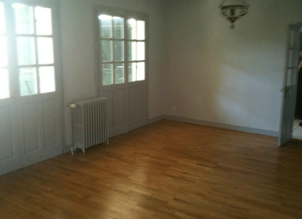 Pièce vide lumineuse avec parquet, deux portes-fenêtres, un radiateur et un lustre au plafond. Murs clairs, porte entrouverte.