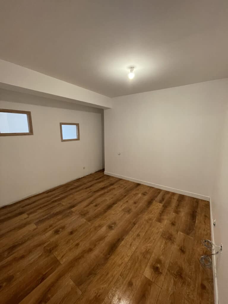 Pièce vide rénovée : murs clairs, parquet moderne, prête à vivre. Pièce vide récemment rénovée avec murs clairs, parquet flottant neuf, deux petites fenêtres encadrées de bois, et fils électriques apparents.