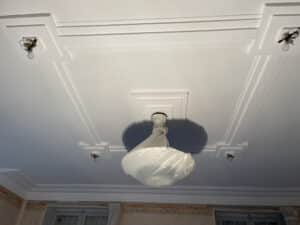Plafond blanc fraîchement peint avec moulures décoratives complexes. Un lustre central sous plastique et quatre ampoules nues sont installés.