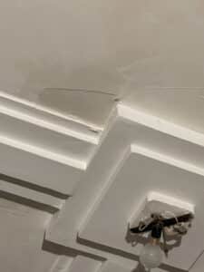Plafond beige avec moulures blanches, présentant des fissures et de la peinture écaillée. Une ampoule pend d'un support d'éclairage provisoire.