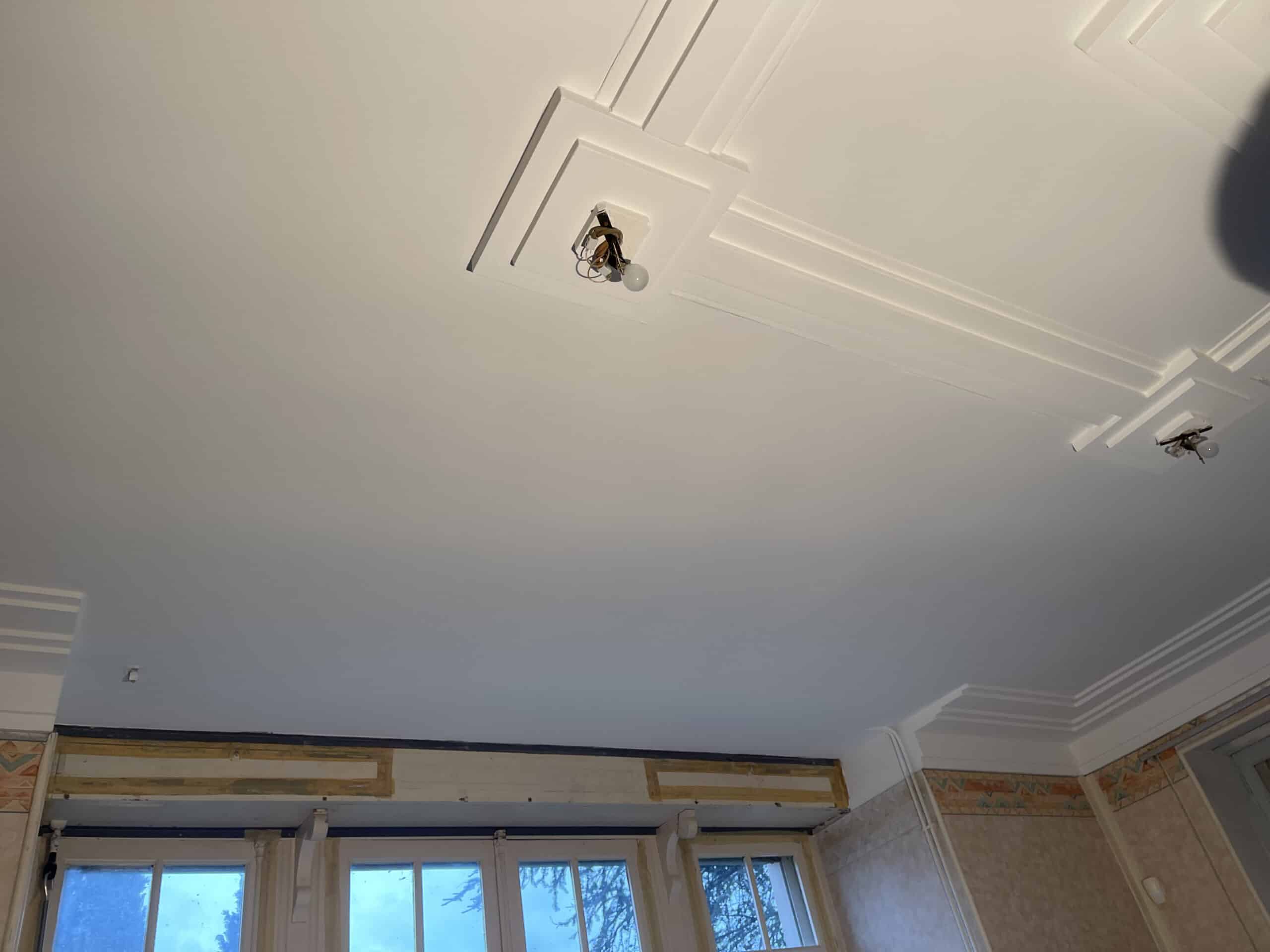 Plafond blanc fraîchement peint avec moulures géométriques et deux luminaires bruts. Le bas montre des fenêtres et des murs en rénovation.