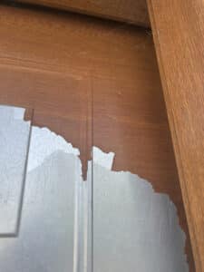 Porte en bois marron dont le placage s'écaille, révélant une surface gris clair rayée et poussiéreuse. Toiles d'araignées visibles.