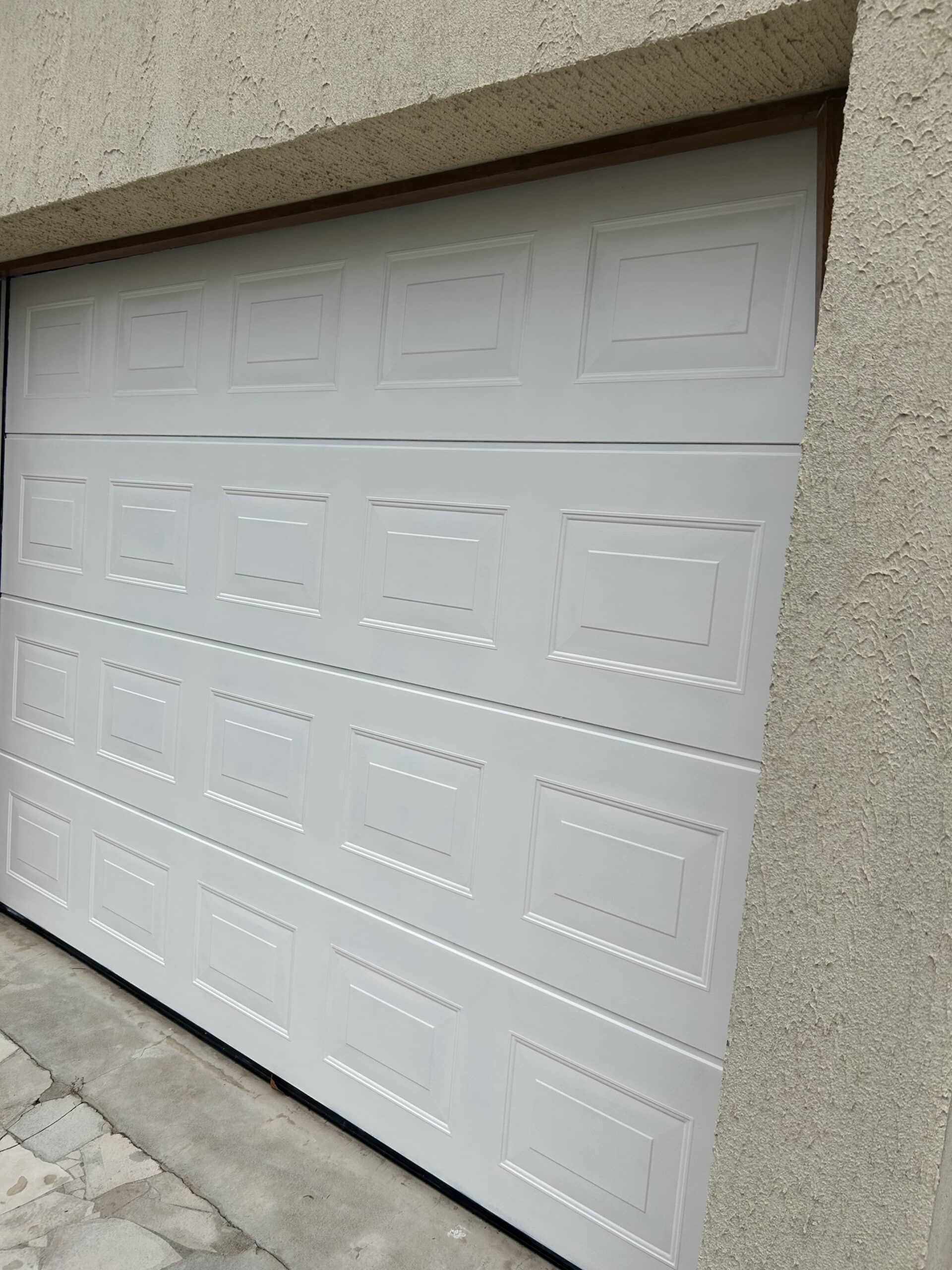 Porte de garage sectionnelle blanche à panneaux fermée, mur crépi beige, sol en béton et dalles. Installation moderne.