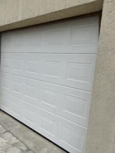 Porte de garage sectionnelle blanche moderne avec panneaux texturés encadrée par une garniture en bois foncé sur un mur de stuc beige, avec un sol pavé.