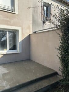 Façade de maison beige et grise avec fenêtres, volet roulant, garde-corps. Patio en béton avec escalier et caniveau, sous le soleil.