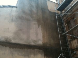 Façade de bâtiment en rénovation avec échafaudage, présentant des zones nettoyées et sales. Des traces d'eau marquent la différence.