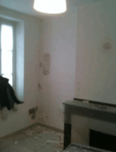 Pièce en rénovation : murs au plâtre écaillé, grande fenêtre lumineuse, cheminée. Sol jonché de débris de travaux.