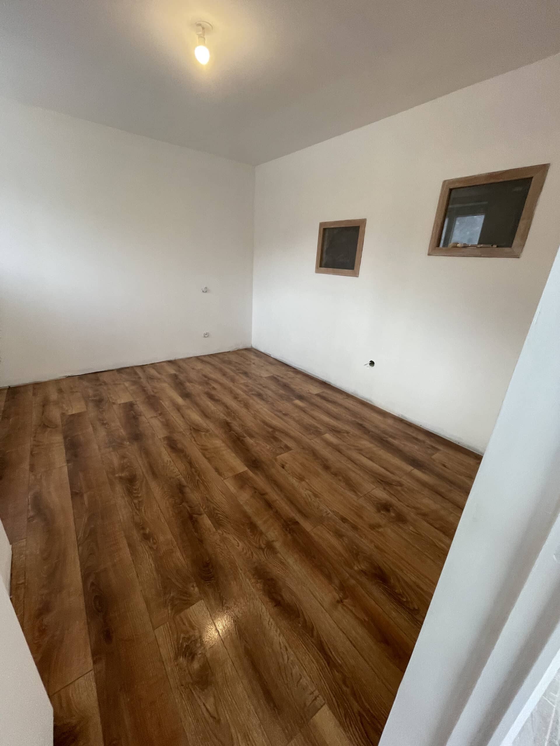 Pièce vide lumineuse avec parquet flottant clair et murs blancs. Une ampoule éclaire les deux ouvertures encadrées en bois et les prises murales.