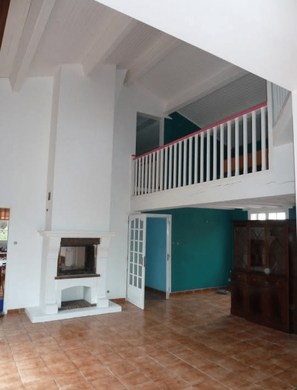 Intérieur de maison avec salon à cheminée blanche, carrelage terracotta. Mezzanine à balustrade blanche et rose surplombant murs bleu sarcelle.