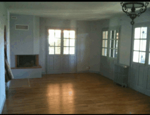 Grand salon vide avec parquet clair, cheminée grise, portes-fenêtres lumineuses et lustre. La lumière naturelle se reflète au sol.