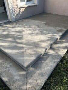 Terrasse en béton à deux niveaux avec finition texturée, bordant un mur de maison et fenêtre. Caniveau de drainage, ombres d'arbres.