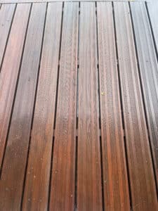 Lames de terrasse en bois rougeâtre, mouillées et rainurées, avec des débris éparpillés. Surface brillante et antidérapante.