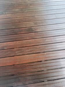 Terrasse en bois humide, lames striées marron foncé avec reflets, posées horizontalement, quelques débris visibles.