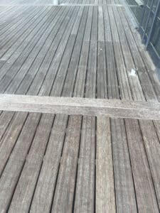 Terrasse en bois gris patiné, lattes rainurées, avec une bordure claire. Petit luminaire et balustrade en verre moderne sur le côté.